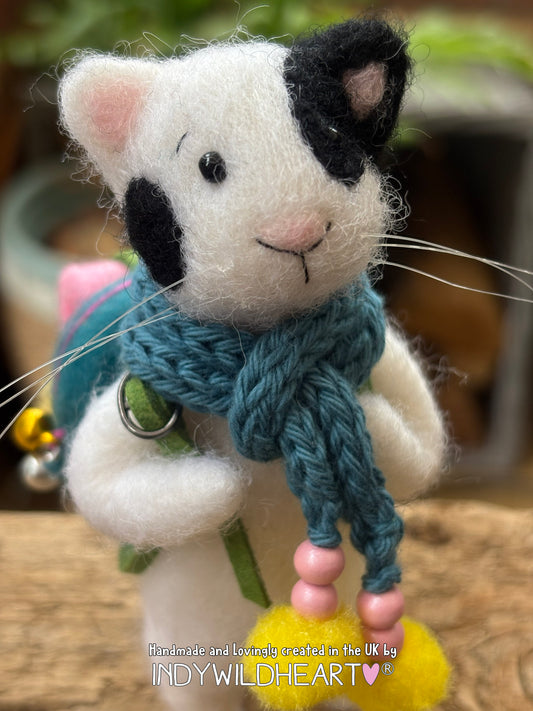 A handmade needle felted wool cat with black and white fur, wearing a blue knit scarf with pink and yellow pom-pom, photographed close-up against a blurred background.