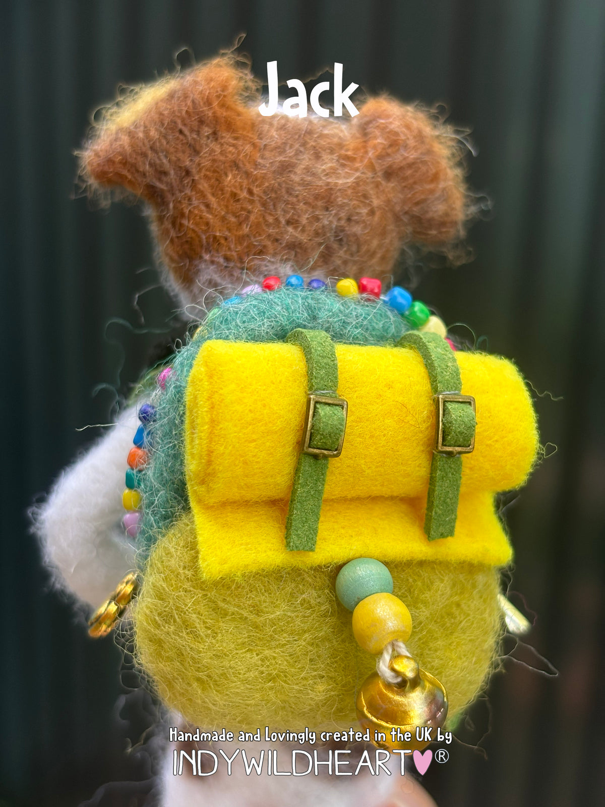 A close-up of a handmade felt dog backpack accessory with a brown felt dog head on top, a bright yellow front flap, green straps, colorful bead accents, and a small gold bell. The word 'Jack' is overlaid at the top, and a watermark at the bottom reads 'Handmade and lovingly created in the UK by INDYWILDEART'.