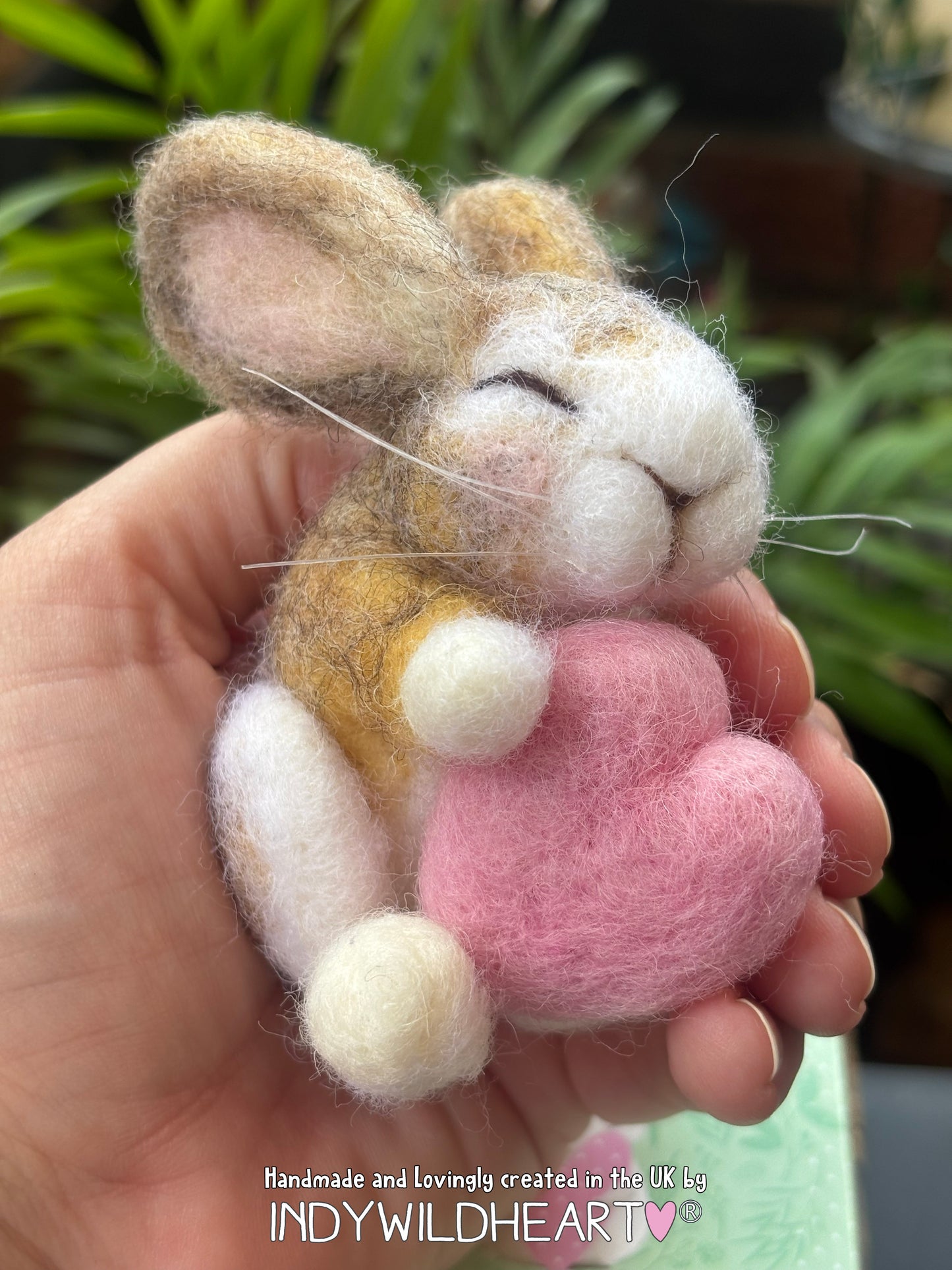 Needle Felt sleeping rabbit holding a little pink heart is being held in the palm of a hand, with foliage behind