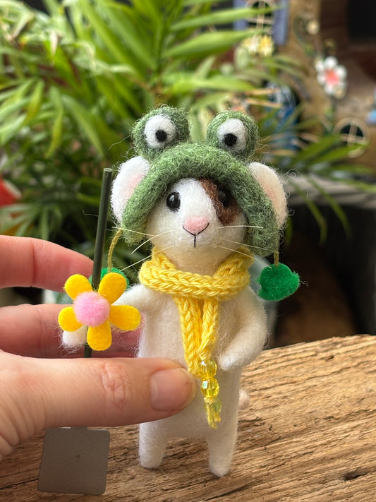 a white felted mouse with a brown patch over one eye, wearing a green frog hat and yellow knitted scarf, holding a miniature garden spade and yellow flower, with a green foliage background. The mouse is being held between a finger and thumb to show is small size.
