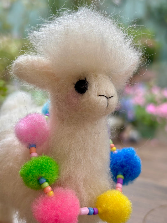 Needle Felted llama with colorful pom-pom accessories against a blurred outdoor background