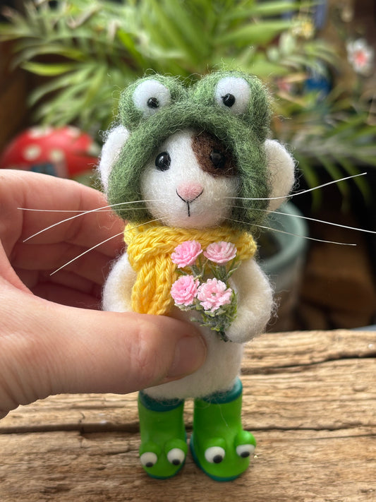 Small needle felted mouse in a frog hat holding flowers, held by a hand on a wooden surface with plants in the background.