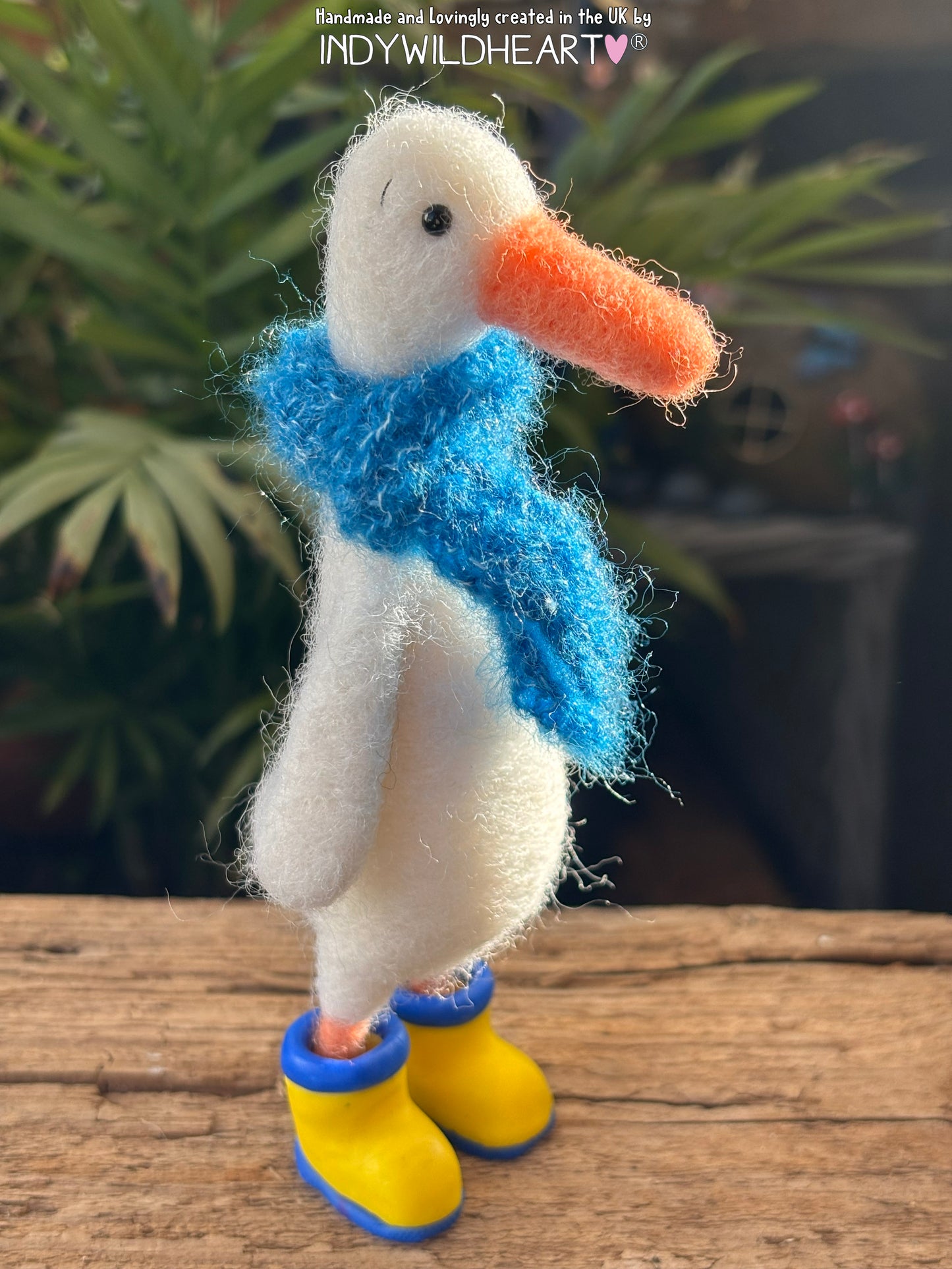 Felted duck with a blue scarf and yellow boots on a wooden surface with plants in the background
