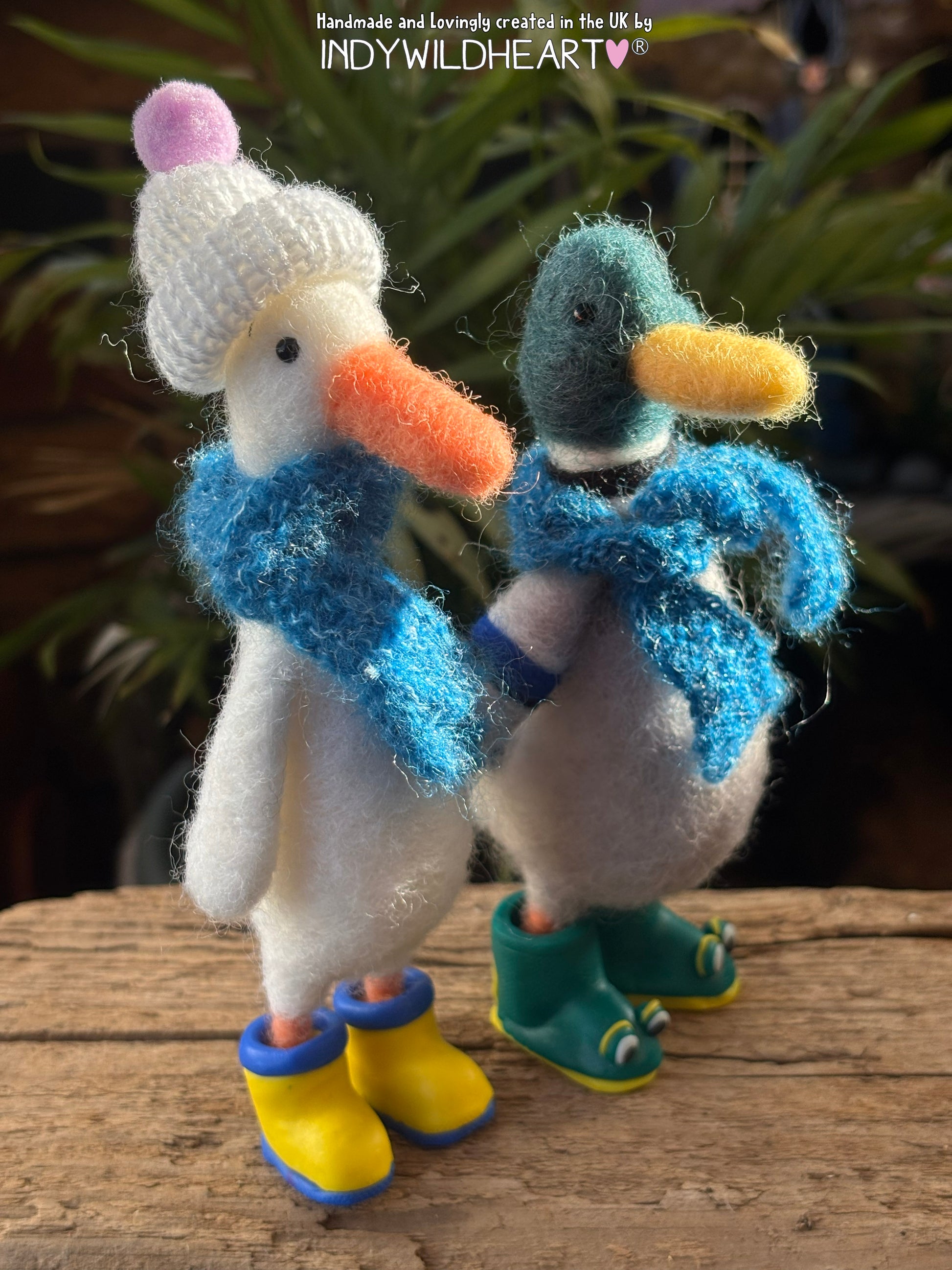 Two felted duck figurines, wearing scarves and boots on a wooden surface with a blurred natural background.