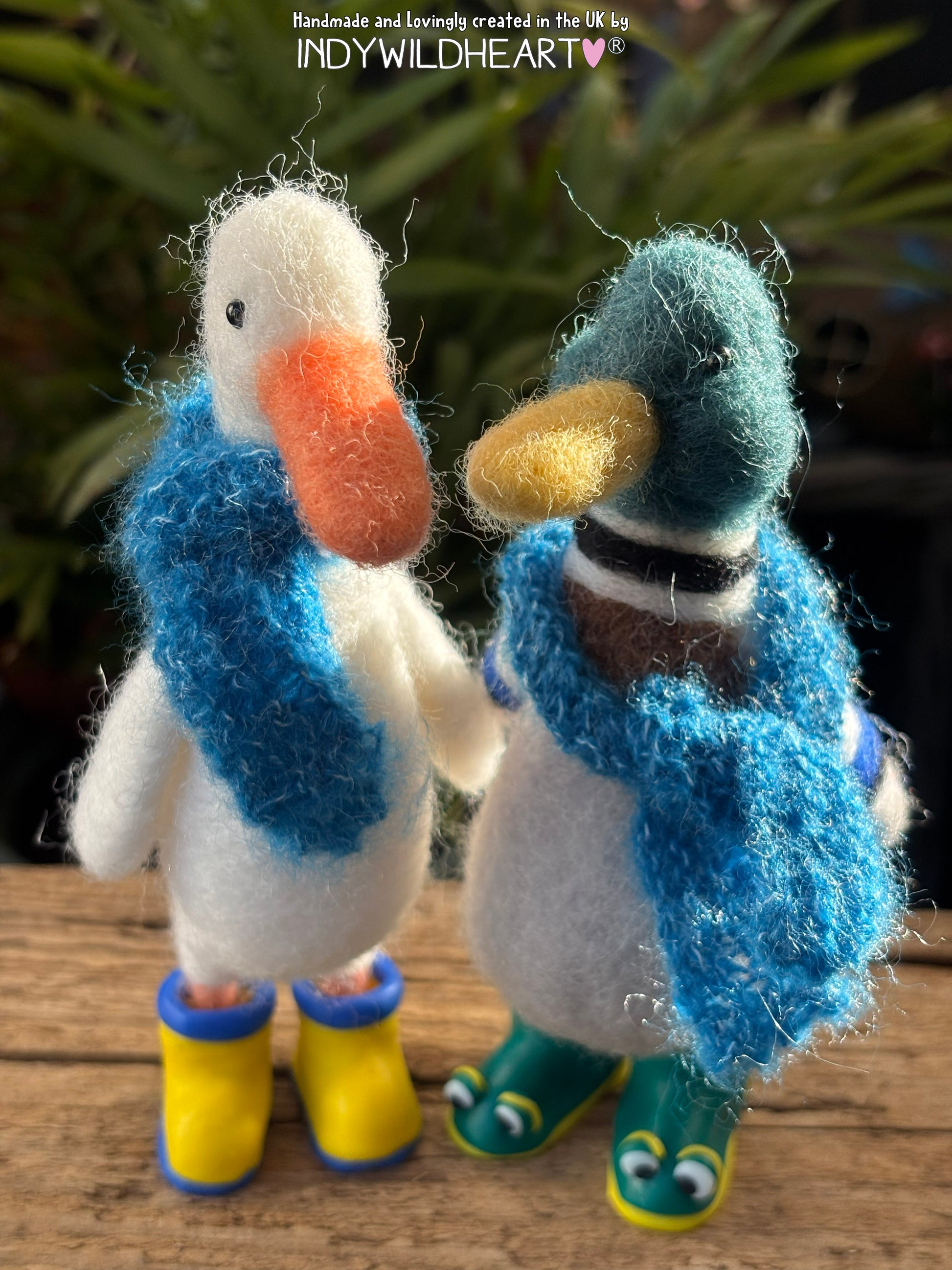 Two felted ducks wearing scarves and boots on a wooden surface with a blurred natural background.