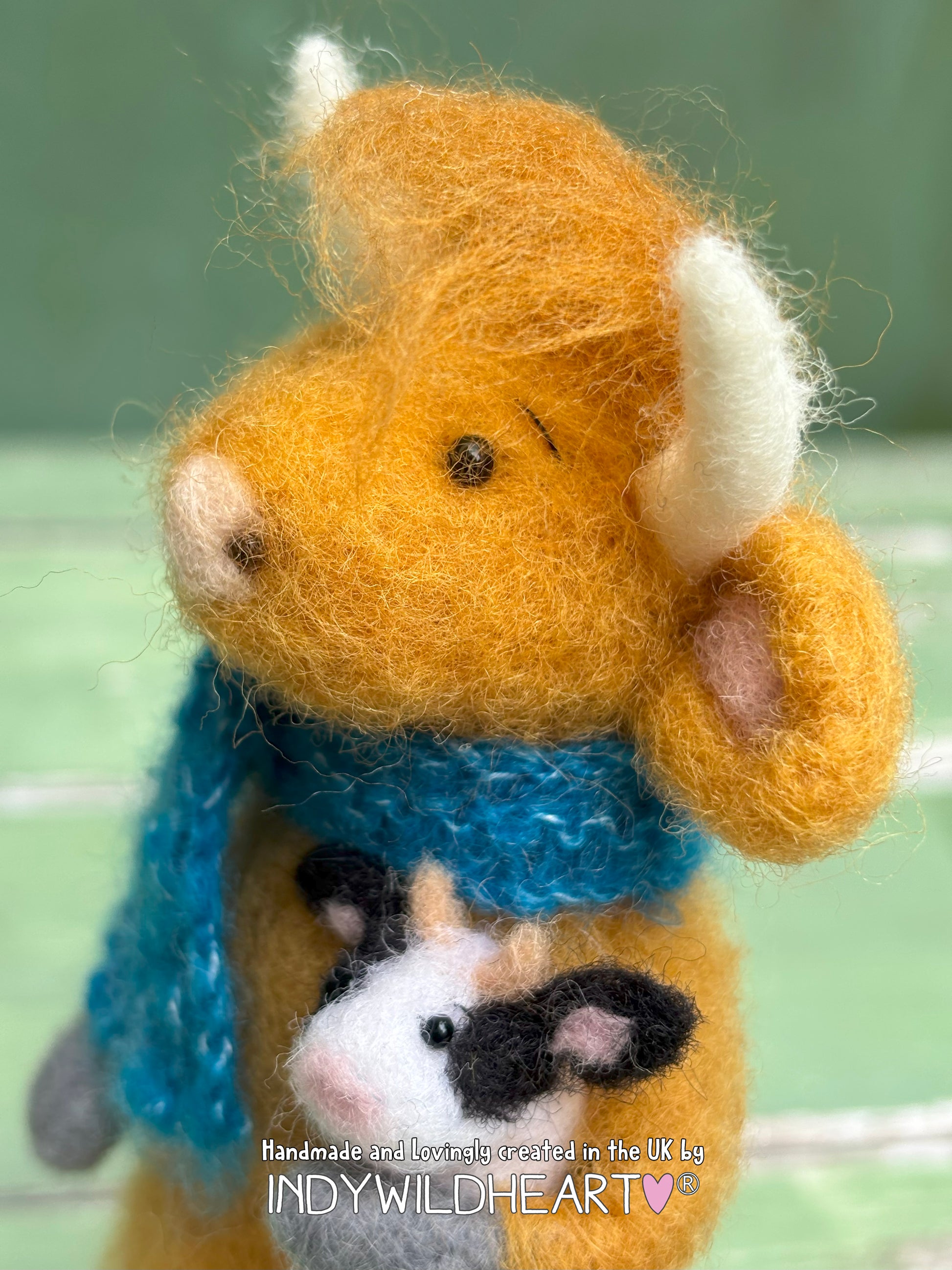 Handmade felted animal a highland cow with a blue scarf and holding a small black and white cow. 