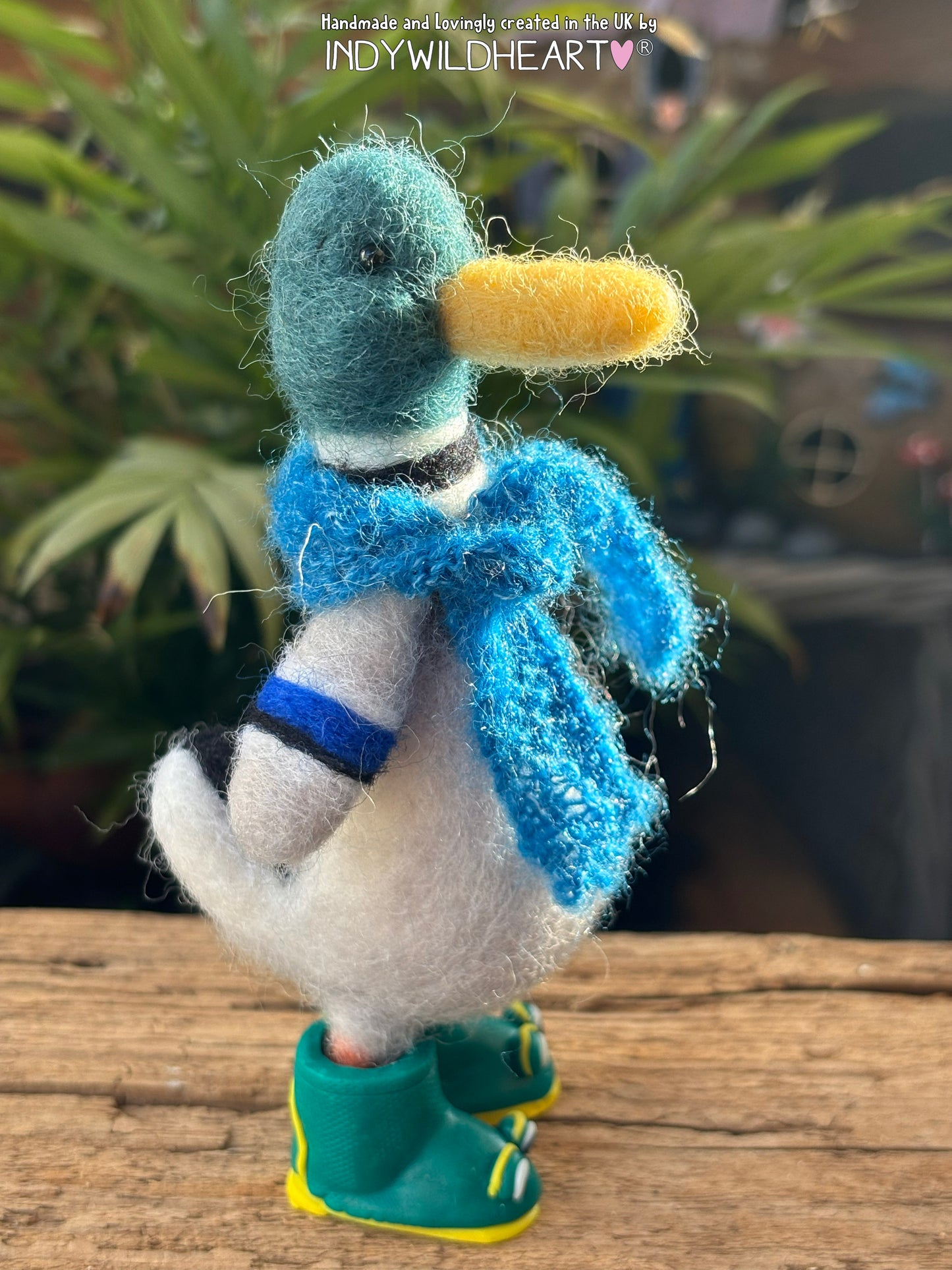 Handmade duck figurine wearing a blue scarf and green boots on a wooden surface with plants in the background.