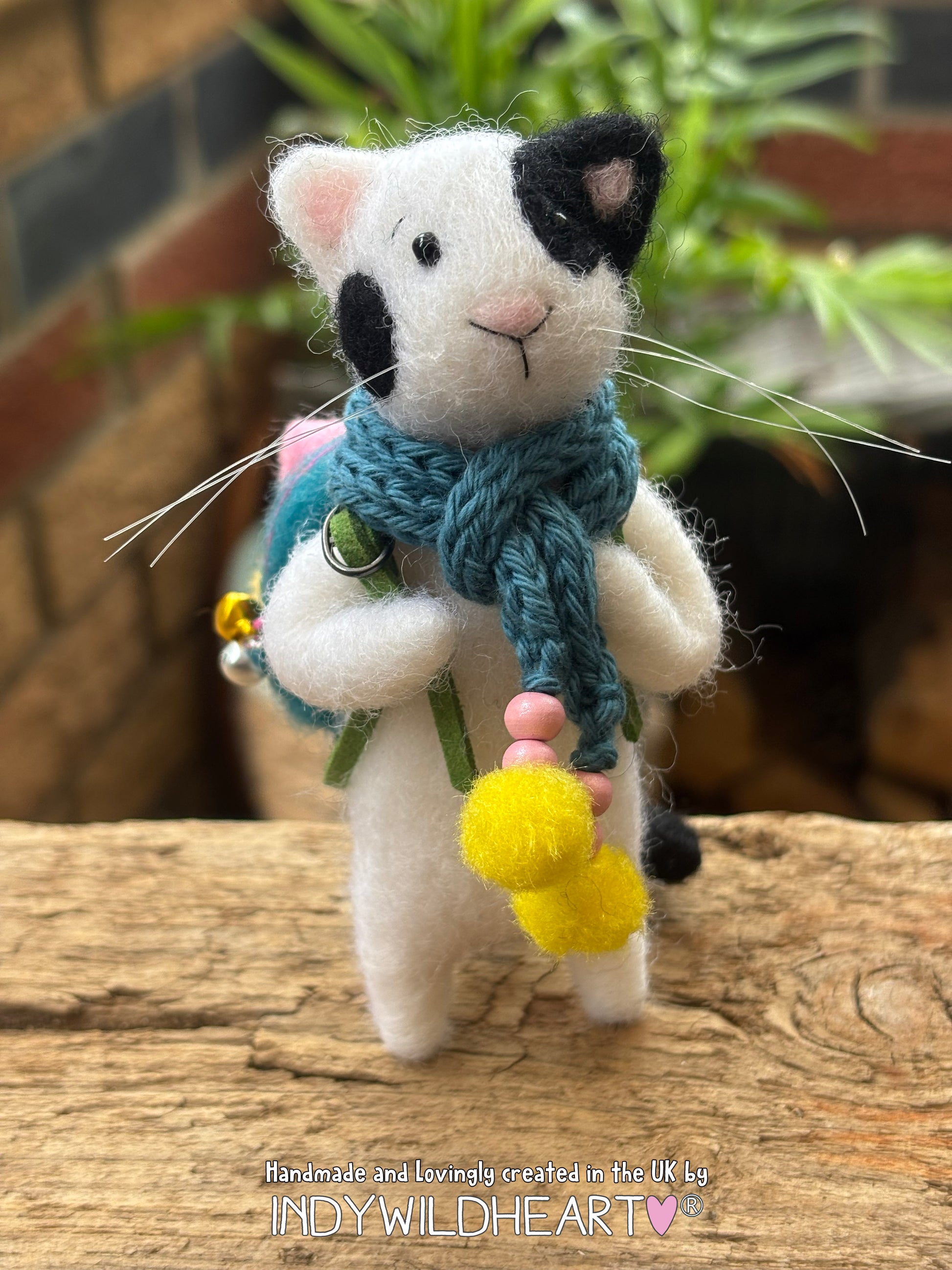 A handmade felted wool cat figurine wearing a blue knit scarf and a small backpack, standing on a rustic wooden surface with a blurred background of plants and a brick wall.