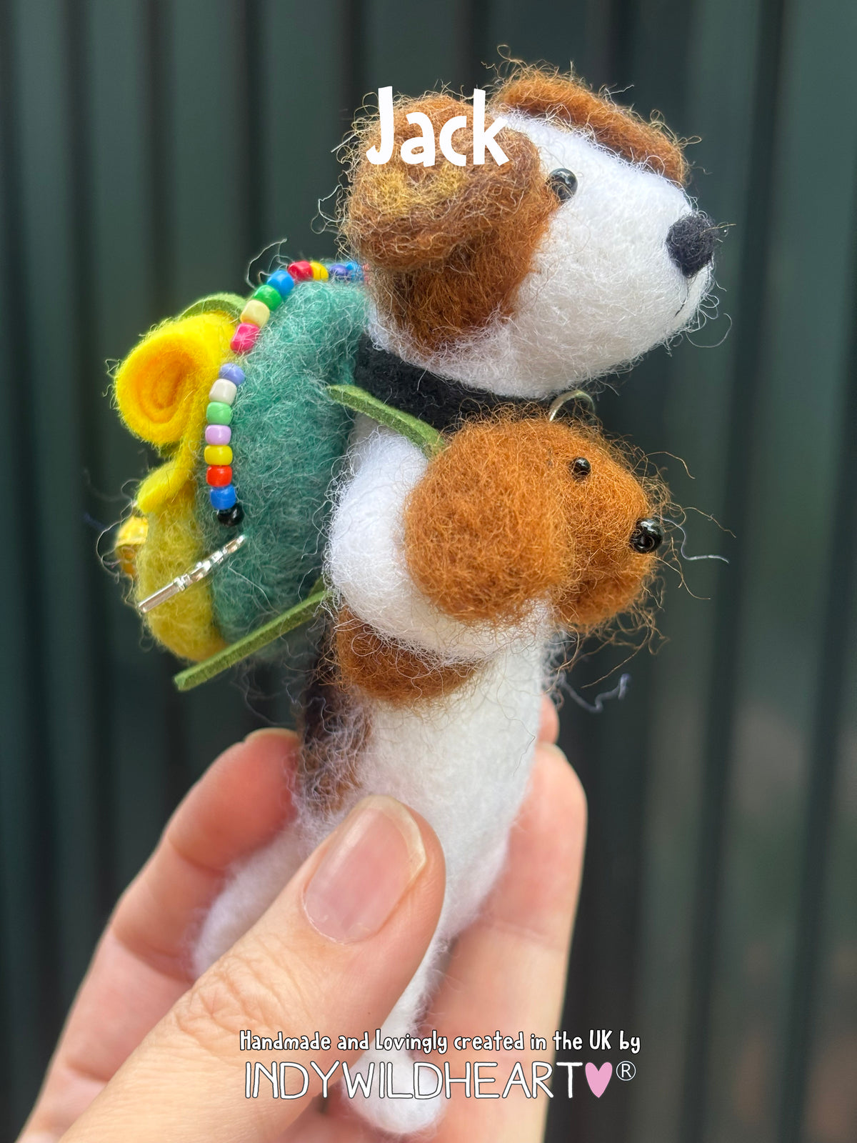 A handmade felted wool dog figurine with a small dog on its back, decorated with a green backpack, yellow flower, and colorful beads. The figurine is held in a person's hand against a dark green slatted background, with the name 'Jack' written above and a brand watermark at the bottom.