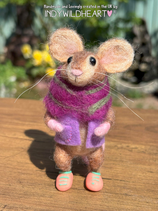 Felted mouse figurine wearing a purple scarf on a wooden surface with blurred greenery in the background.
