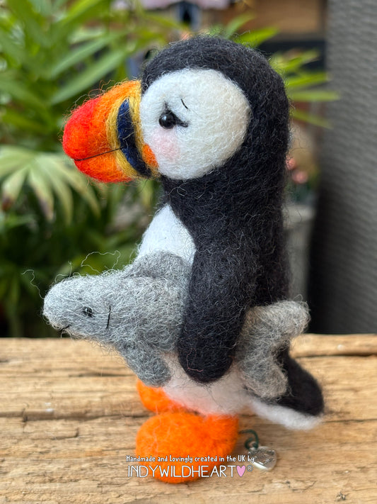 Felted puffin with a fish on a wooden surface, blurred greenery in the background
