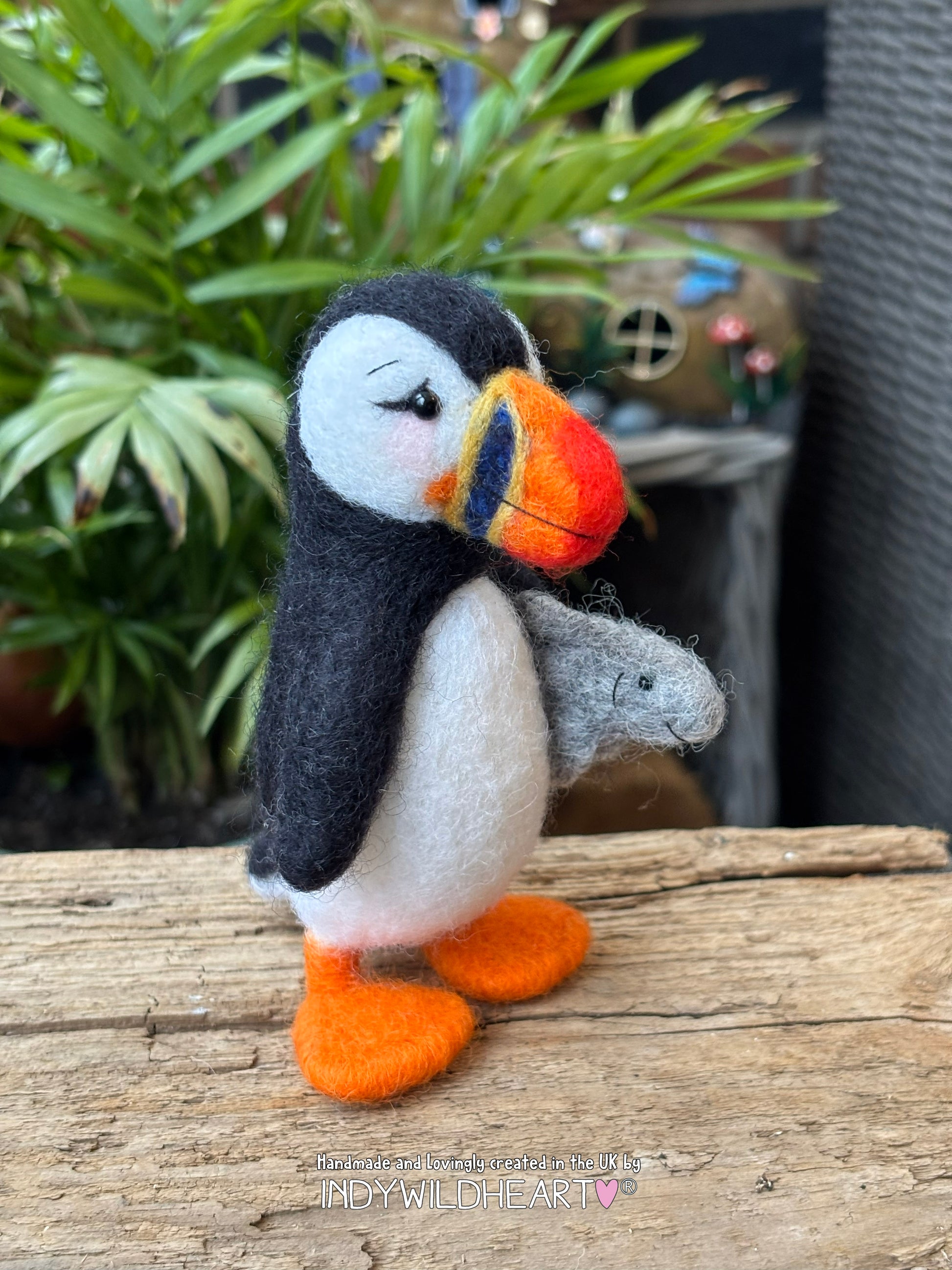 woolly puffin holding a fish on a wooden surface with plants in the background