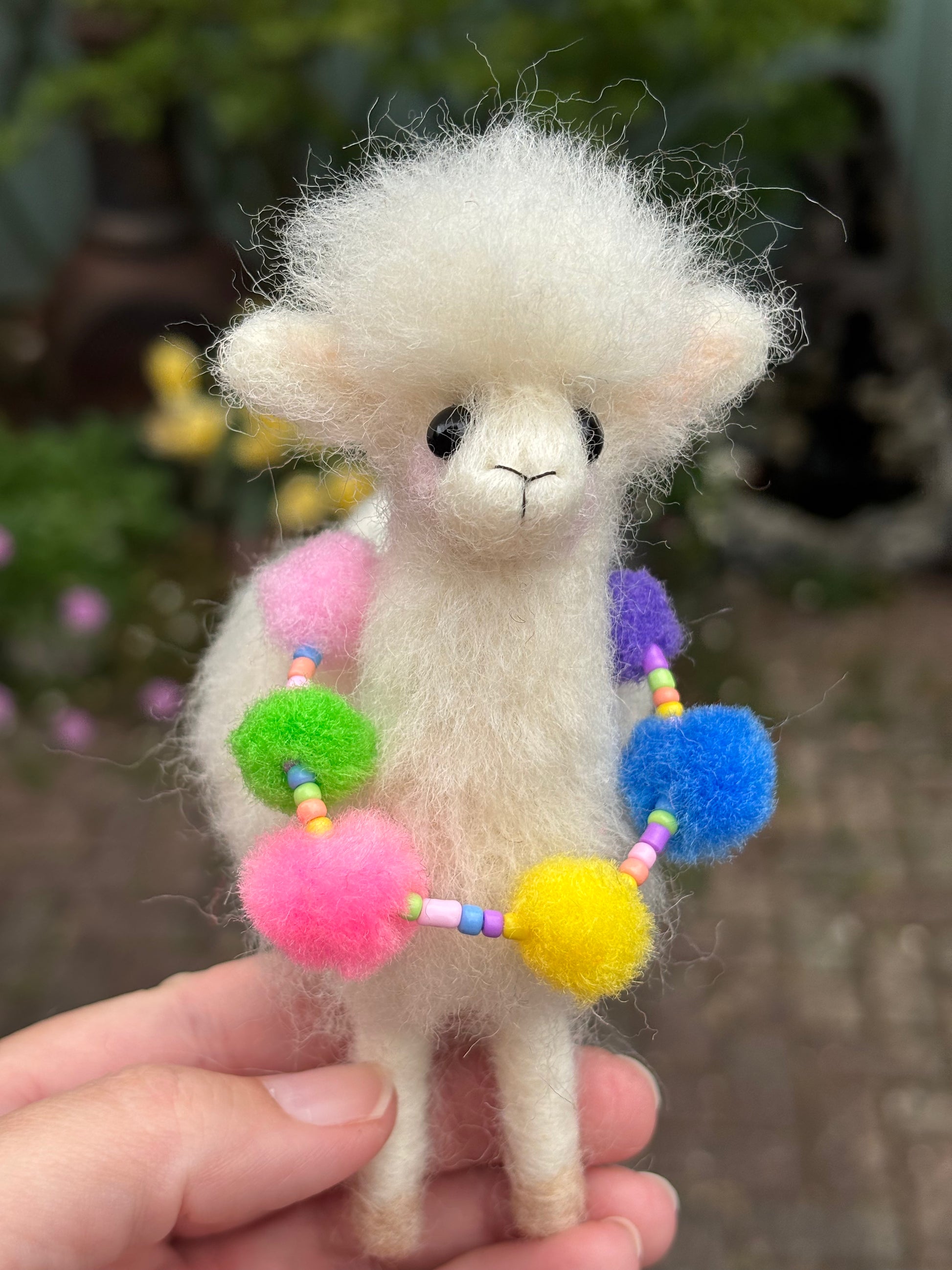 Small fluffy needle felted llama with colorful pom-pom necklace held in a hand against a blurred natural background