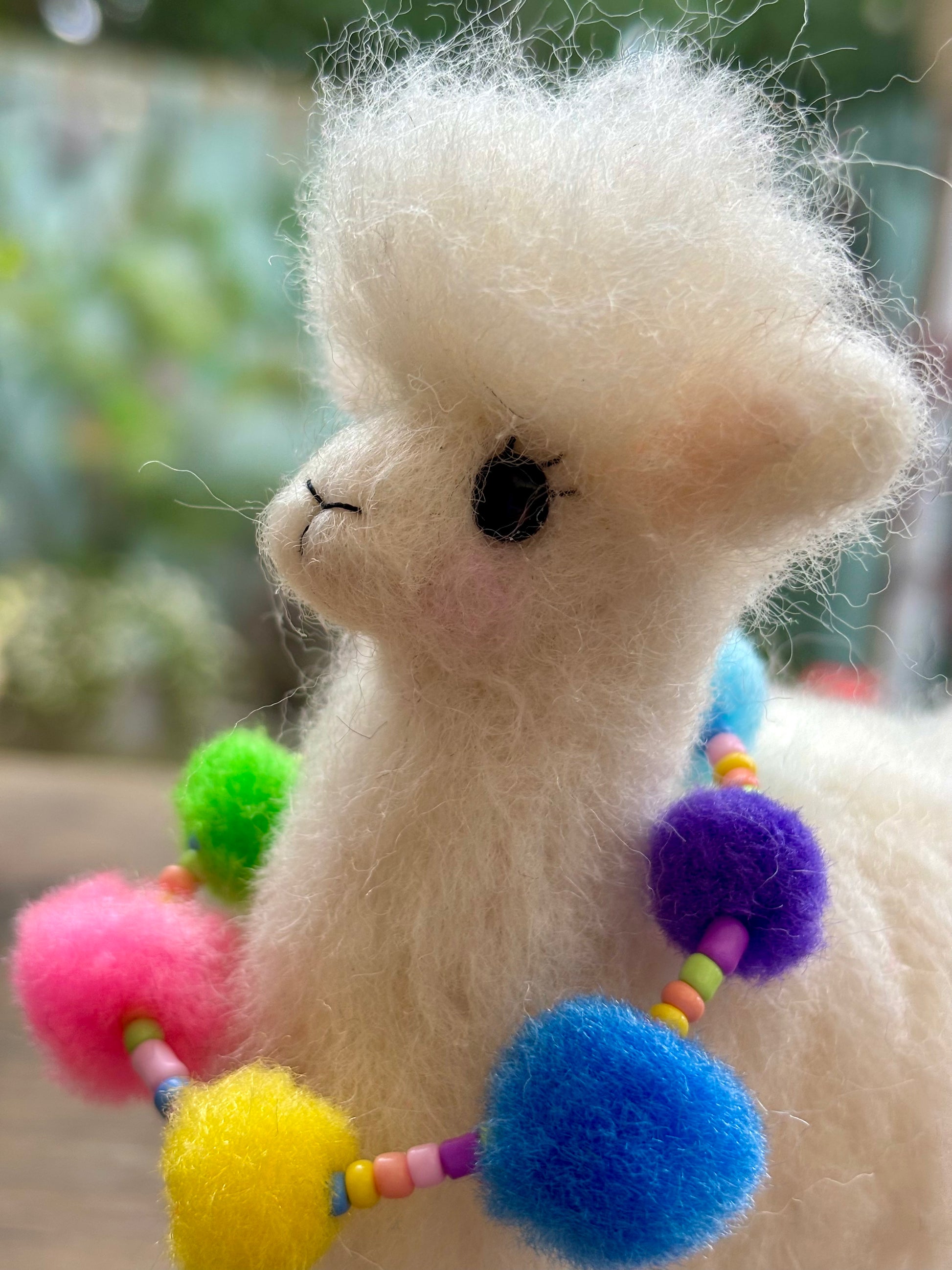White fluffy needle felt Alpaca with colorful pom-pom accessories against a blurred green background