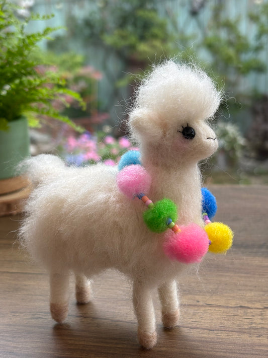 White fluffy needle felted alpaca with colourful pom-pom necklace on a wooden surface with plants in the background