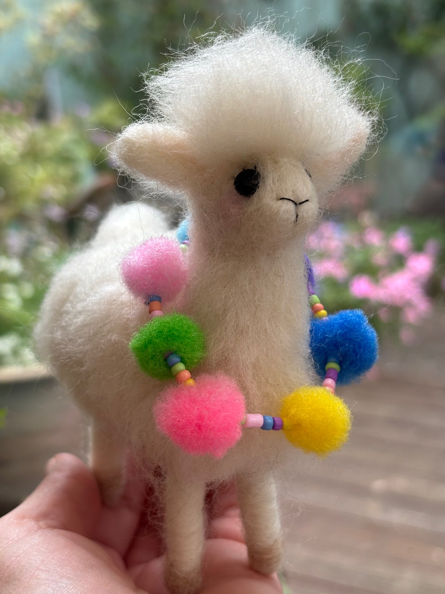 needle felted Alpaca with colorful pom-pom accessories held by a hand against a blurred natural background