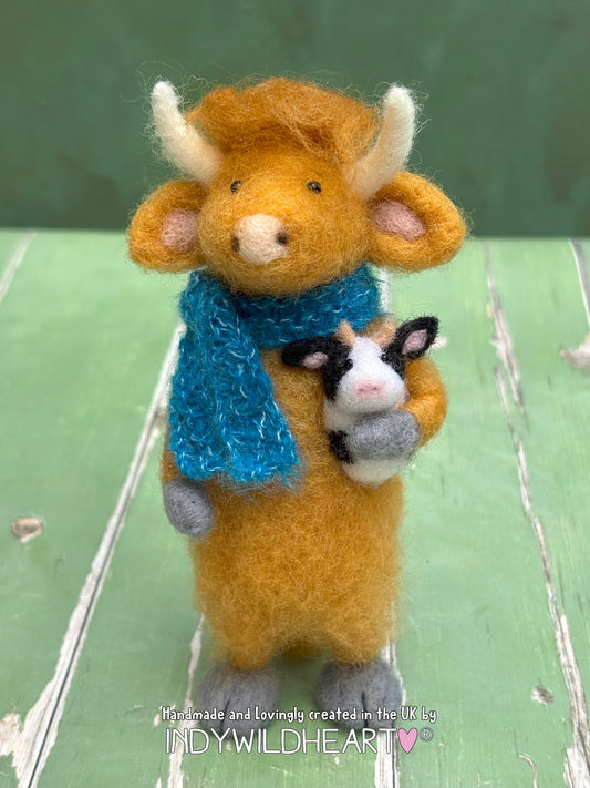 Needle Felted Highland Cow character holding a black and white cow with a blue scarf, on a green background.
