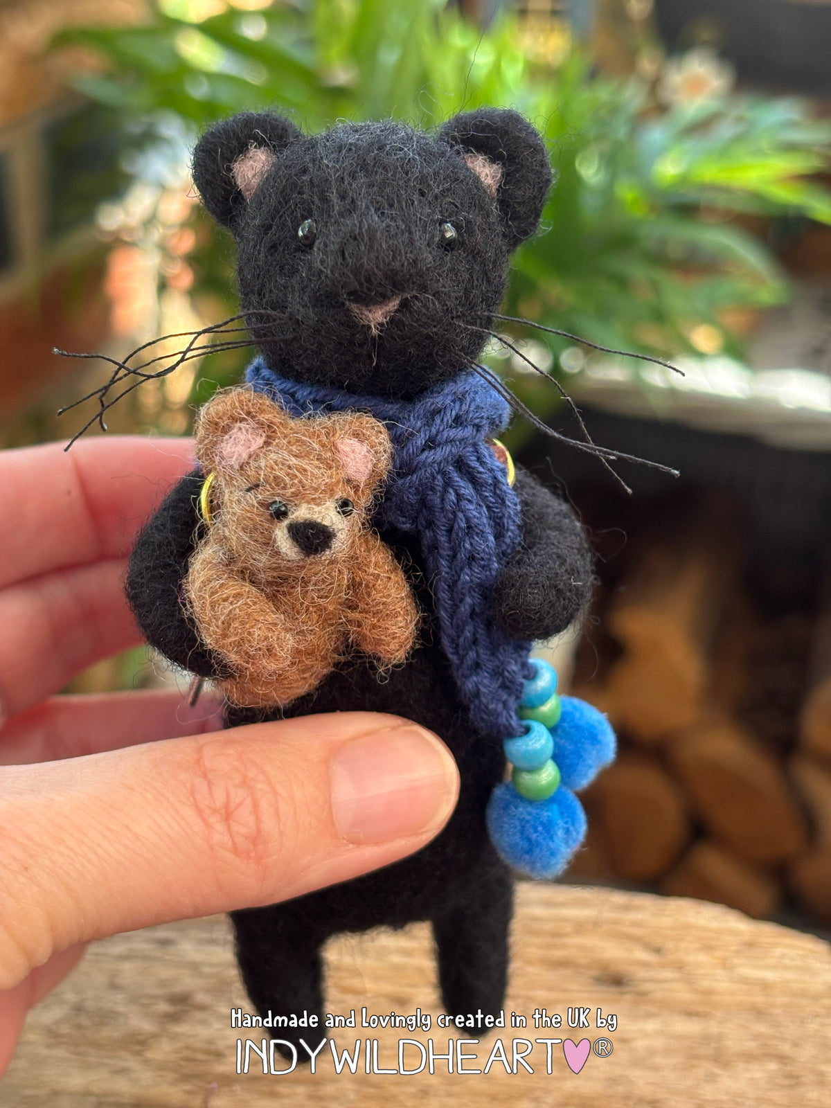 Needle Felted Black Panther character with navy blue scarf and holding a miniature needle felted teddy, being held by a hand to show it's small scale