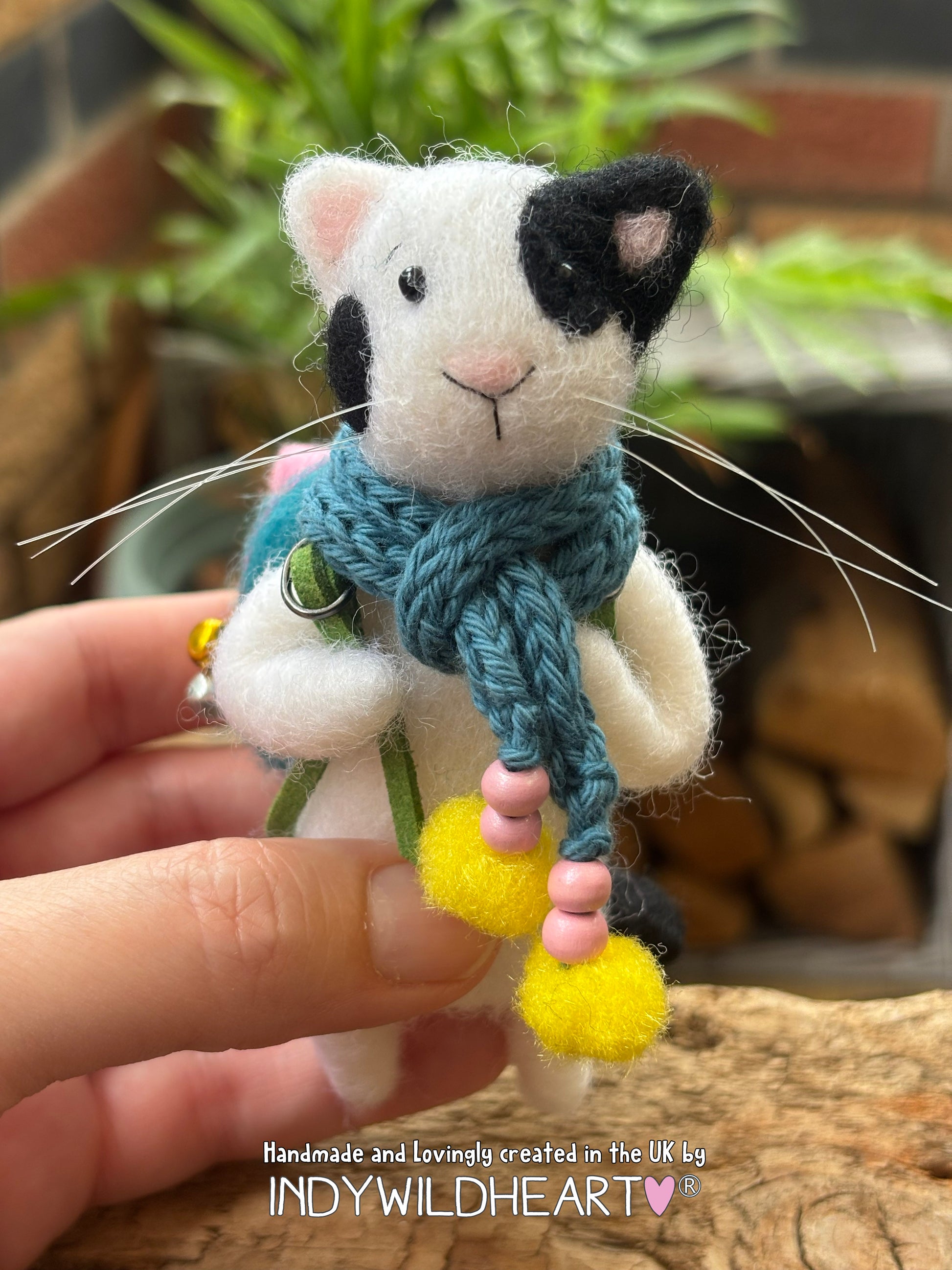 A handmade felted wool cat figurine with a black and white body, a blue knit scarf, and yellow pom-poms, held in a person's hand against a blurred background of plants and wood.