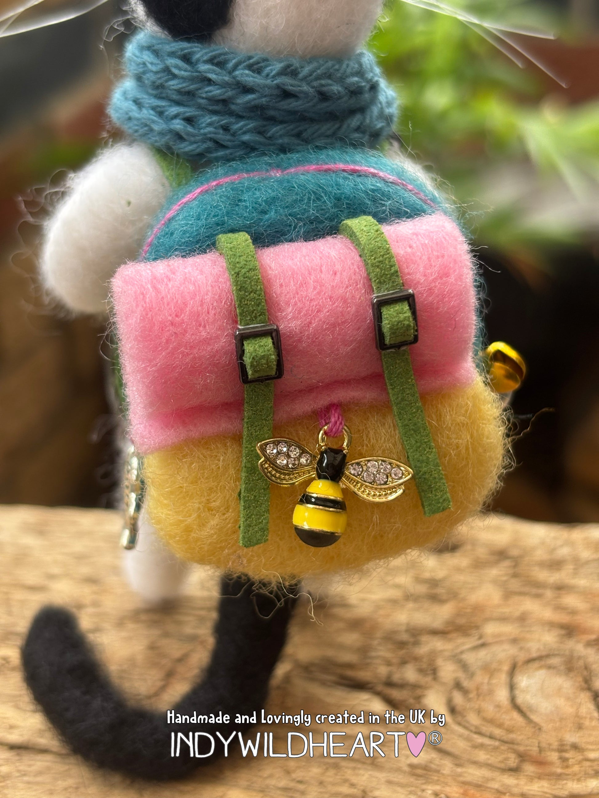 A close-up of a handmade felted wool backpack accessory with a pink top flap, yellow lower body, green straps, and a bumblebee charm, shown attached to a small plush figure on a wooden surface with a blurred outdoor background.