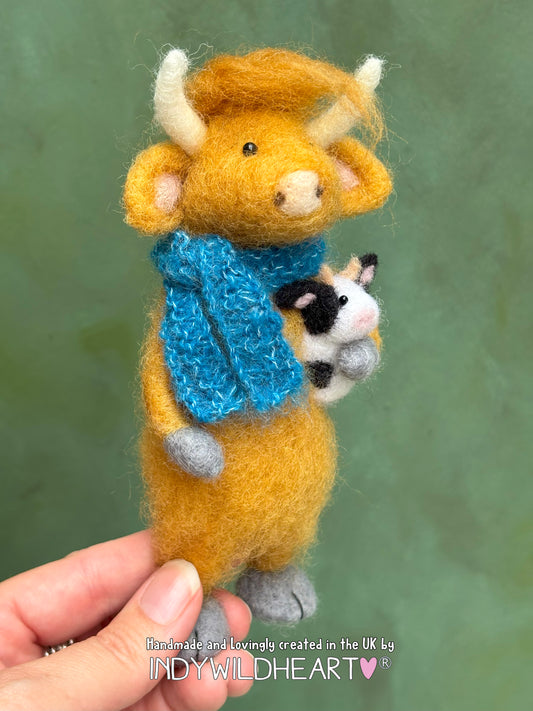 Miniature Felted toy cow holding a teddy cow with a blue scarf, held by a hand against a green background.