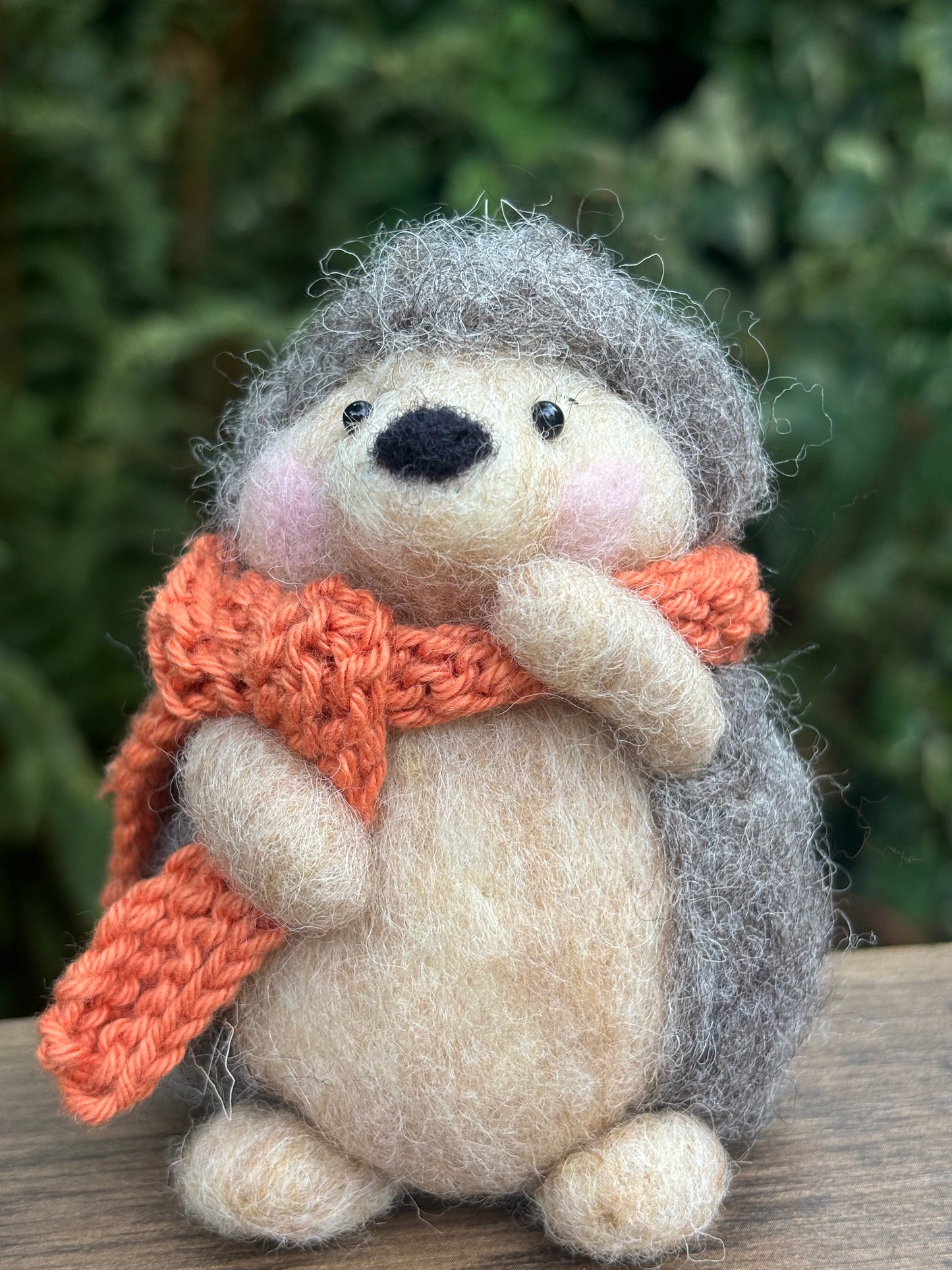 needle felted hedgehog with an orange scarf on a wooden surface with a blurred green background