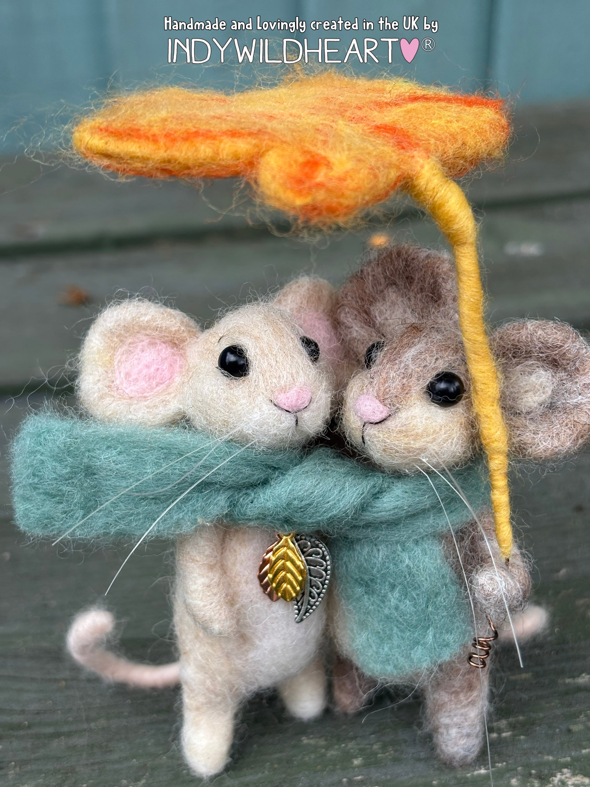 A handmade felted wool art piece depicting two small mice hugging under a yellow leaf-shaped umbrella, wrapped in a teal-green scarf with a gold leaf charm. The image has a text overlay at the top reading 'Handmade and lovingly created in the UK by INDYWILDHART'.