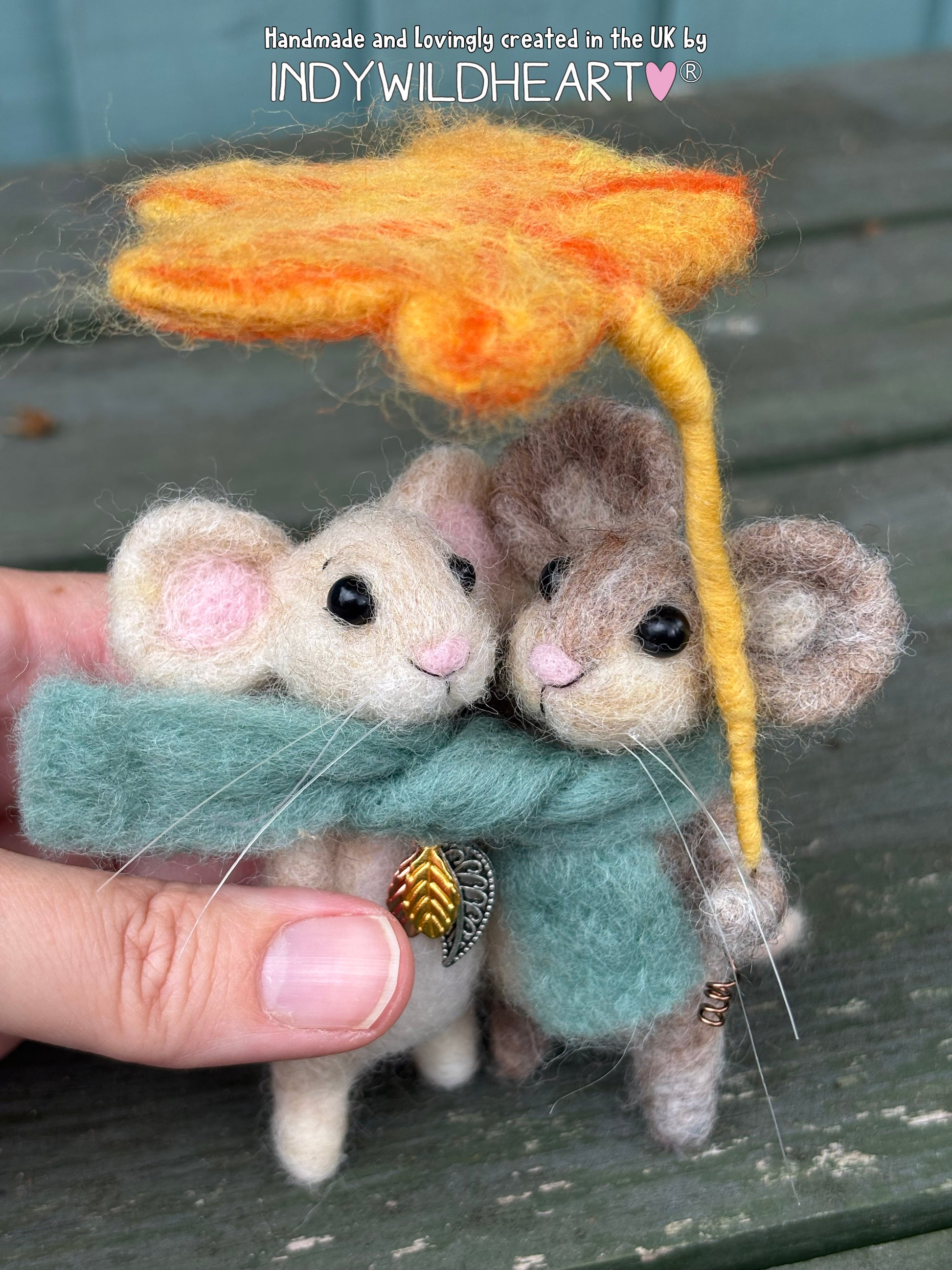 A close-up of a handmade felted wool mouse figurine being held by a hand. The white mouse wears a dusty teal scarf with a small gold chevron charm, while a brown mouse sits beside it under a fuzzy orange-yellow leaf hat.