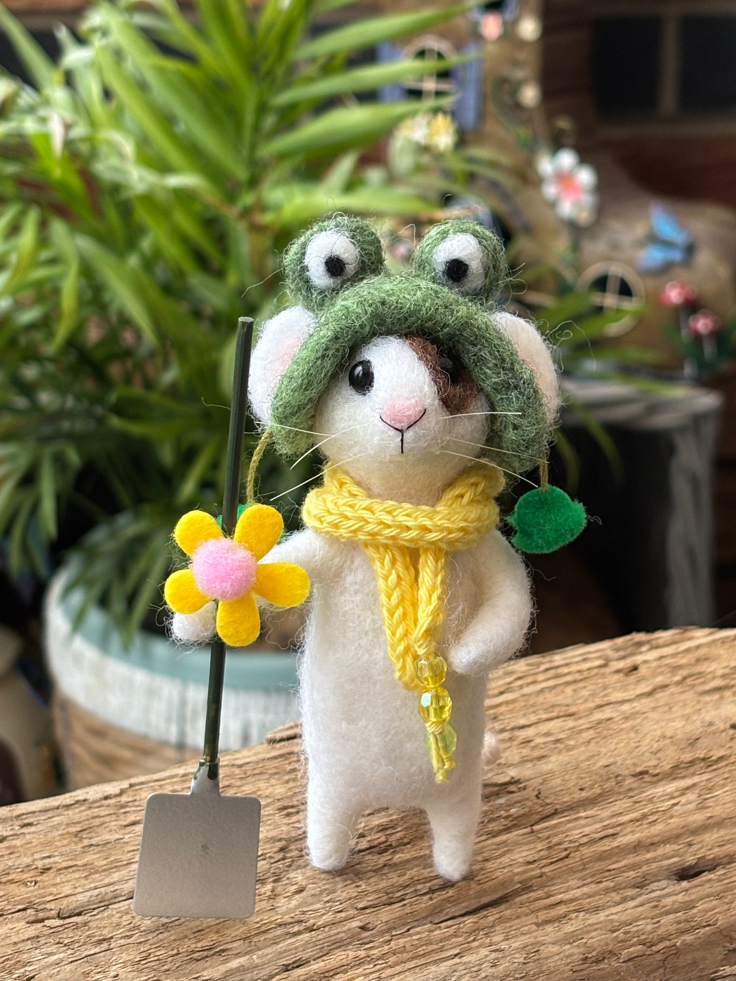 needle felted white mouse with green hat and yellow scarf, holding a miniature garden spade, with green foliage in the background