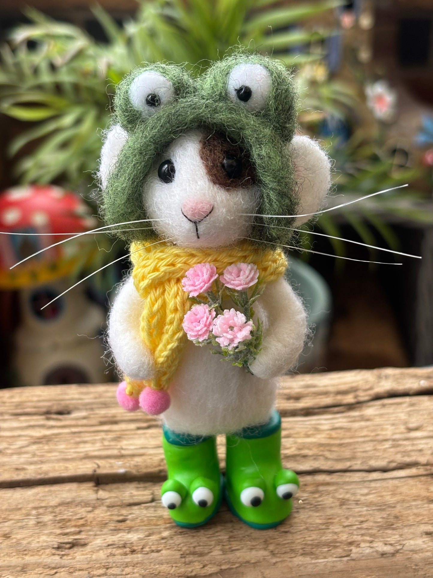 Needle felted white mouse with a brown patch over on eye, wearing a very stylish green frog style hat, and green clay frog wellies. With a yellow scarf, standing on wood with a blurred background.