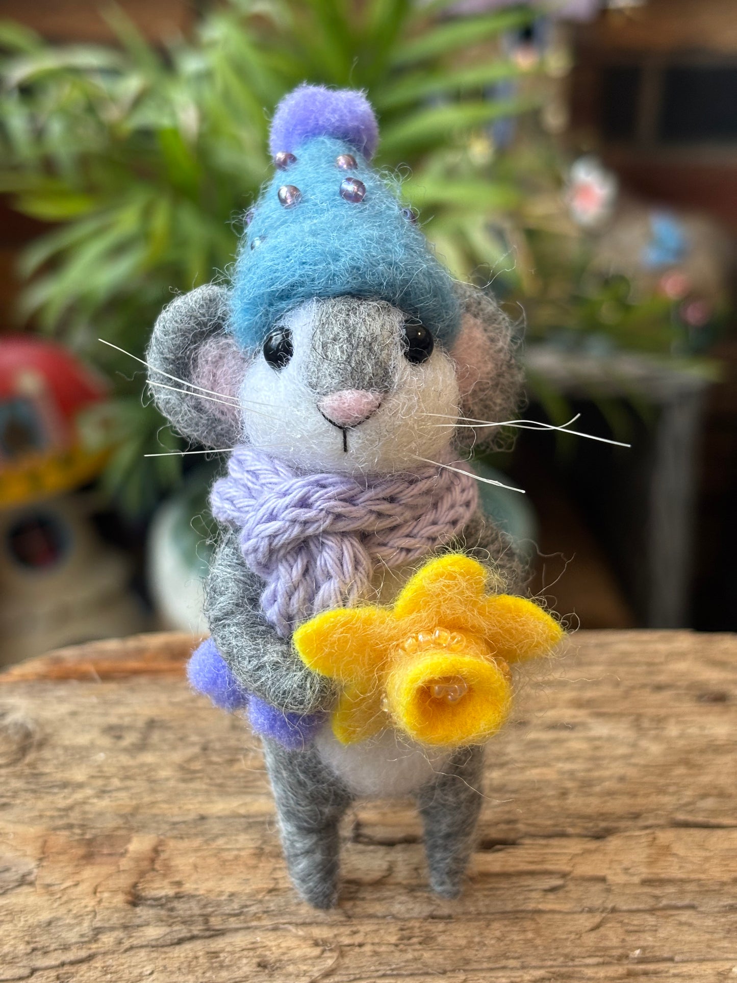 Needle Felted Mouse holding a needle felted daffodil, wearing a lilac hand knitted scarf and light blue bobble hat, on a wood stand with green foliage background