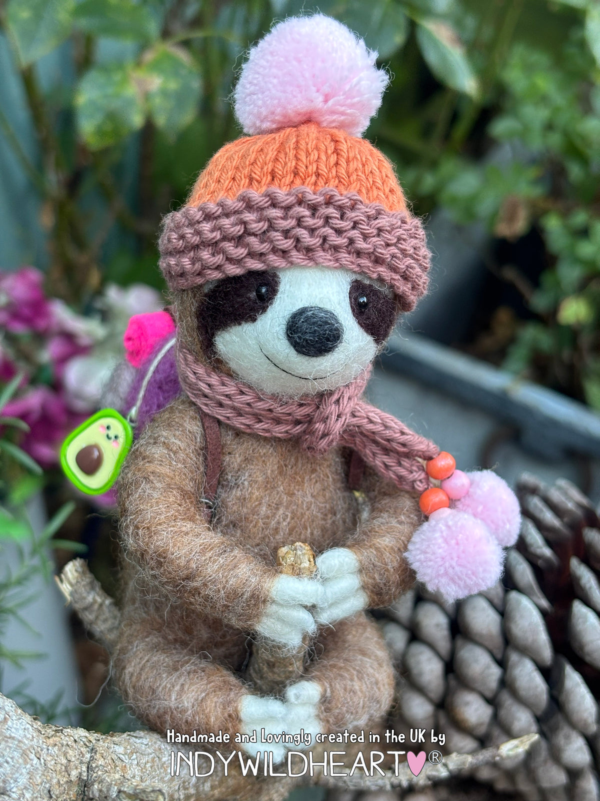 Needle Felted Brown Sloth sitting, wearing a knitted brown & orange hat with a pink bobble.  A brown scarf with pink pom poms and carrying a purple and pink rucksack with miniature hanging charms