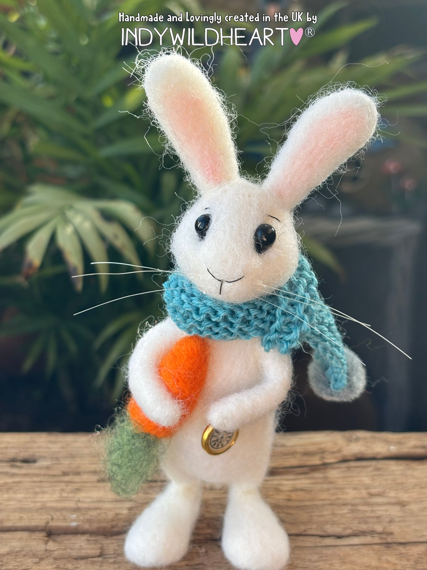 needle felted white rabbit standing with blue knitted scarf and holding a felt carrot and a pocket watch charm with a plant blurred in background