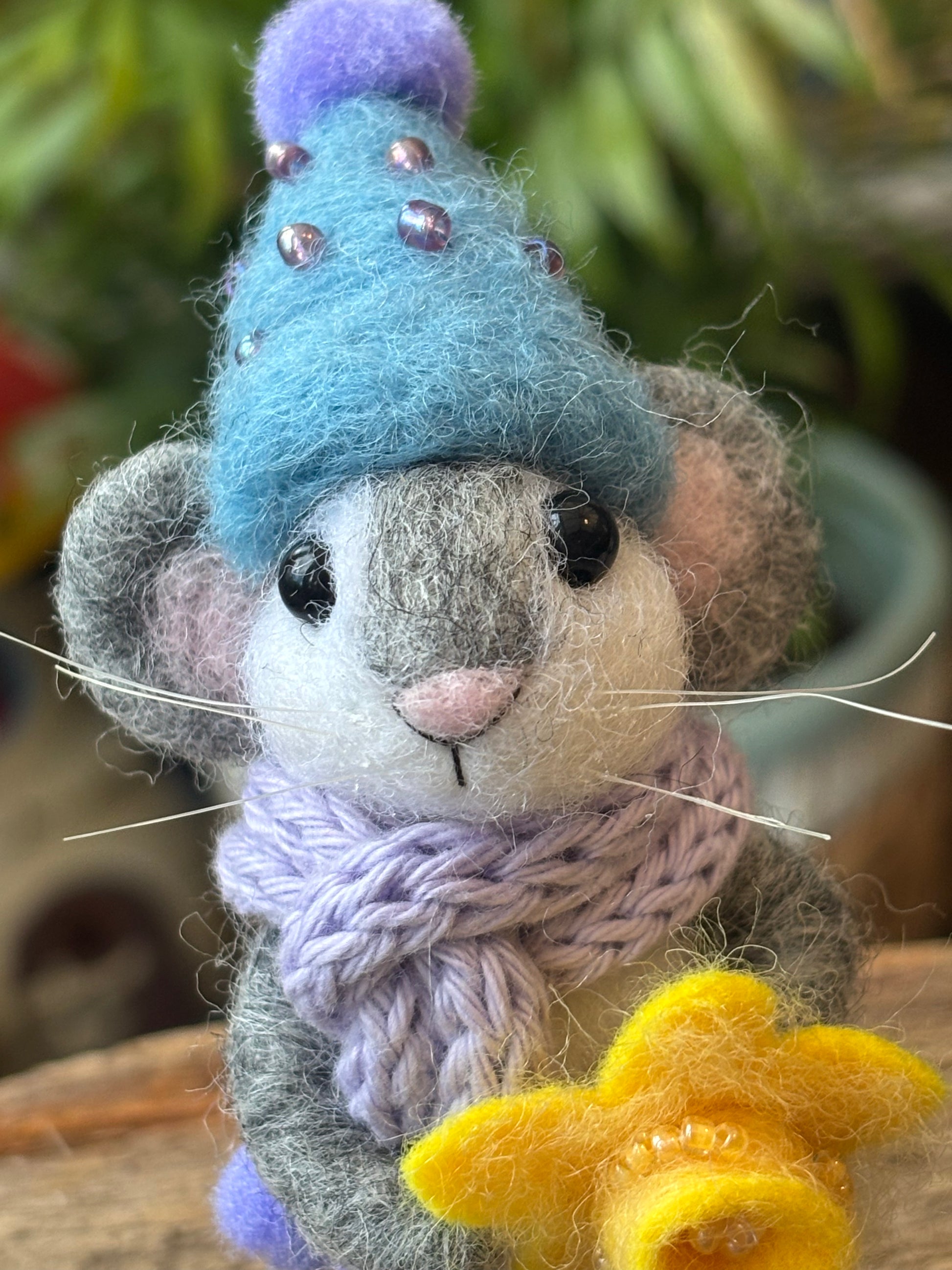 needle felted  mouse wearing a blue hat with purple pom-pom, grey scarf, and holding a yellow daffodil against a blurred green background.