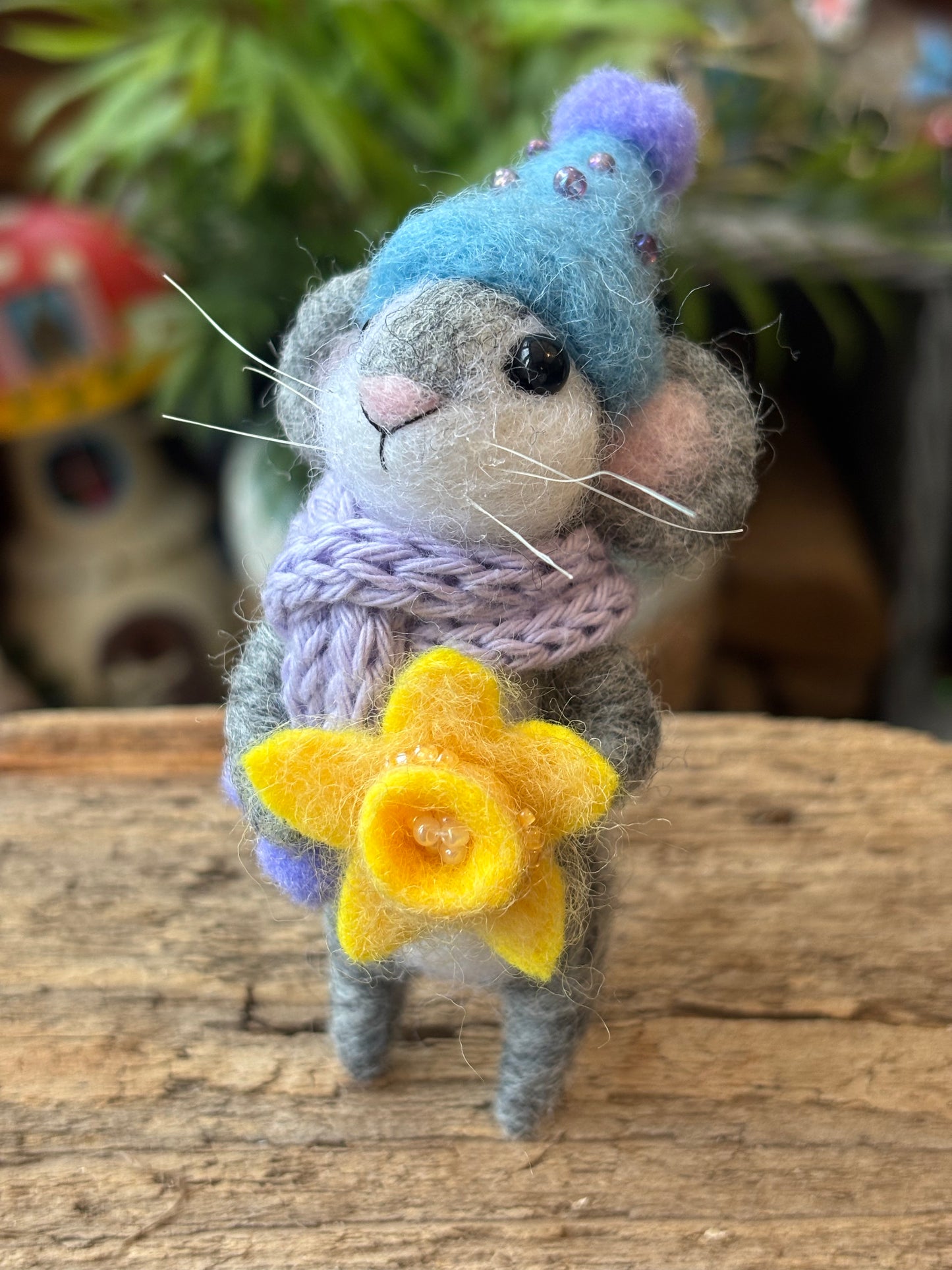 Felted mouse toy with a blue hat, purple scarf, and yellow flower on a wooden surface.