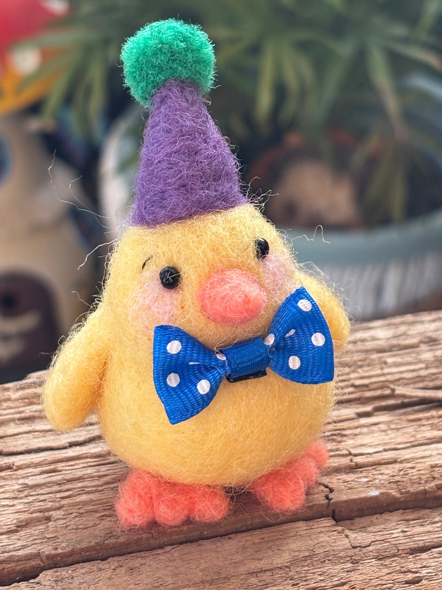 Small yellow felted chick with a purple hat and blue bow tie on a wooden surface.