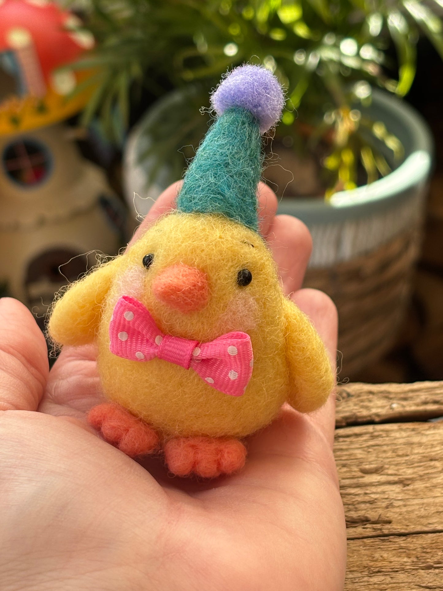 Small yellow felt chick with a colorful hat held in a hand against a natural background.
