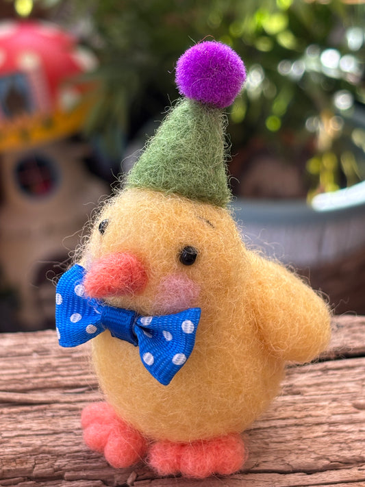 needle felted yellow chick with a green hat and blue bow tie on a wooden surface