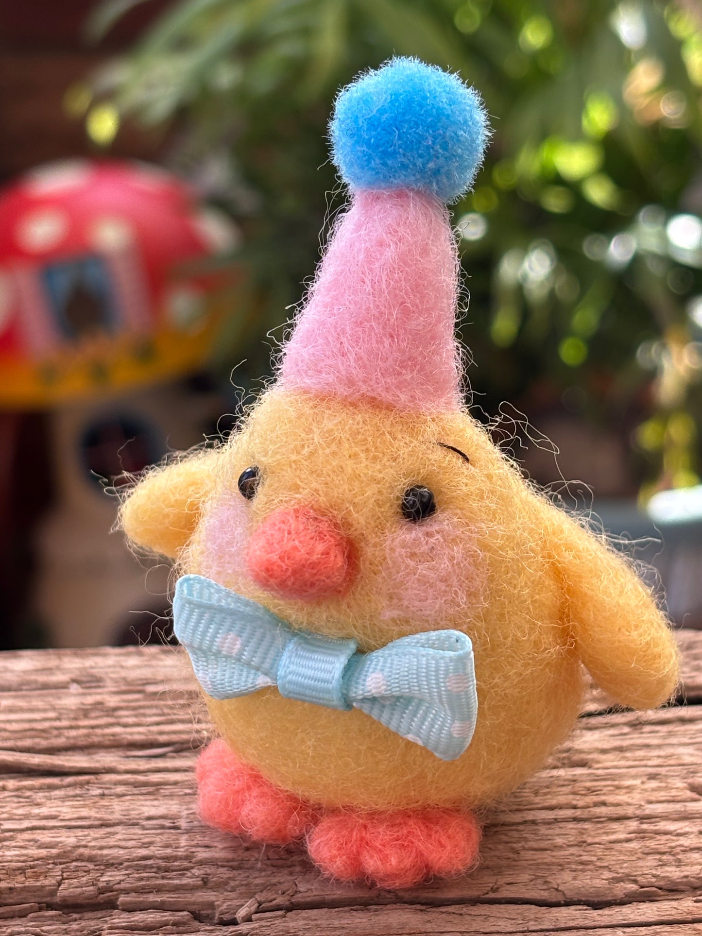 Yellow needle felted chick with a blue bow tie and colorful hat on a wooden surface