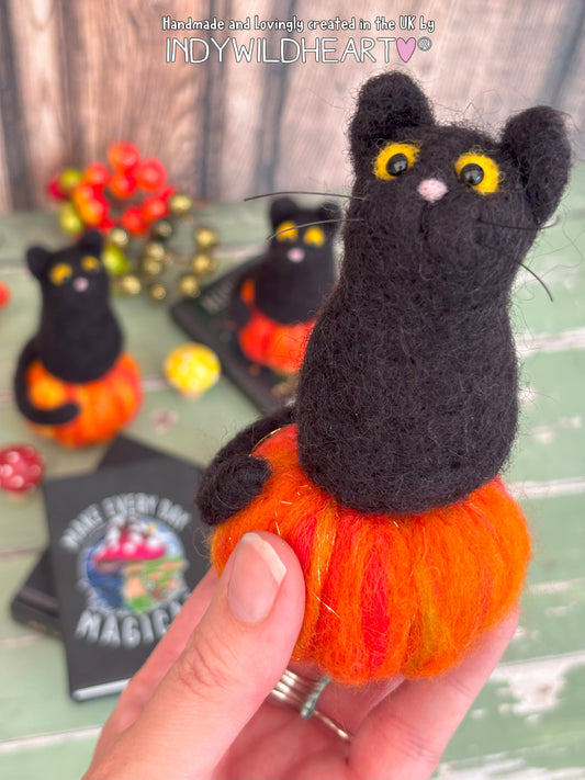 Black cat figurine on an orange pumpkin held by a hand, with 'Indy Wildheart Art' branding.