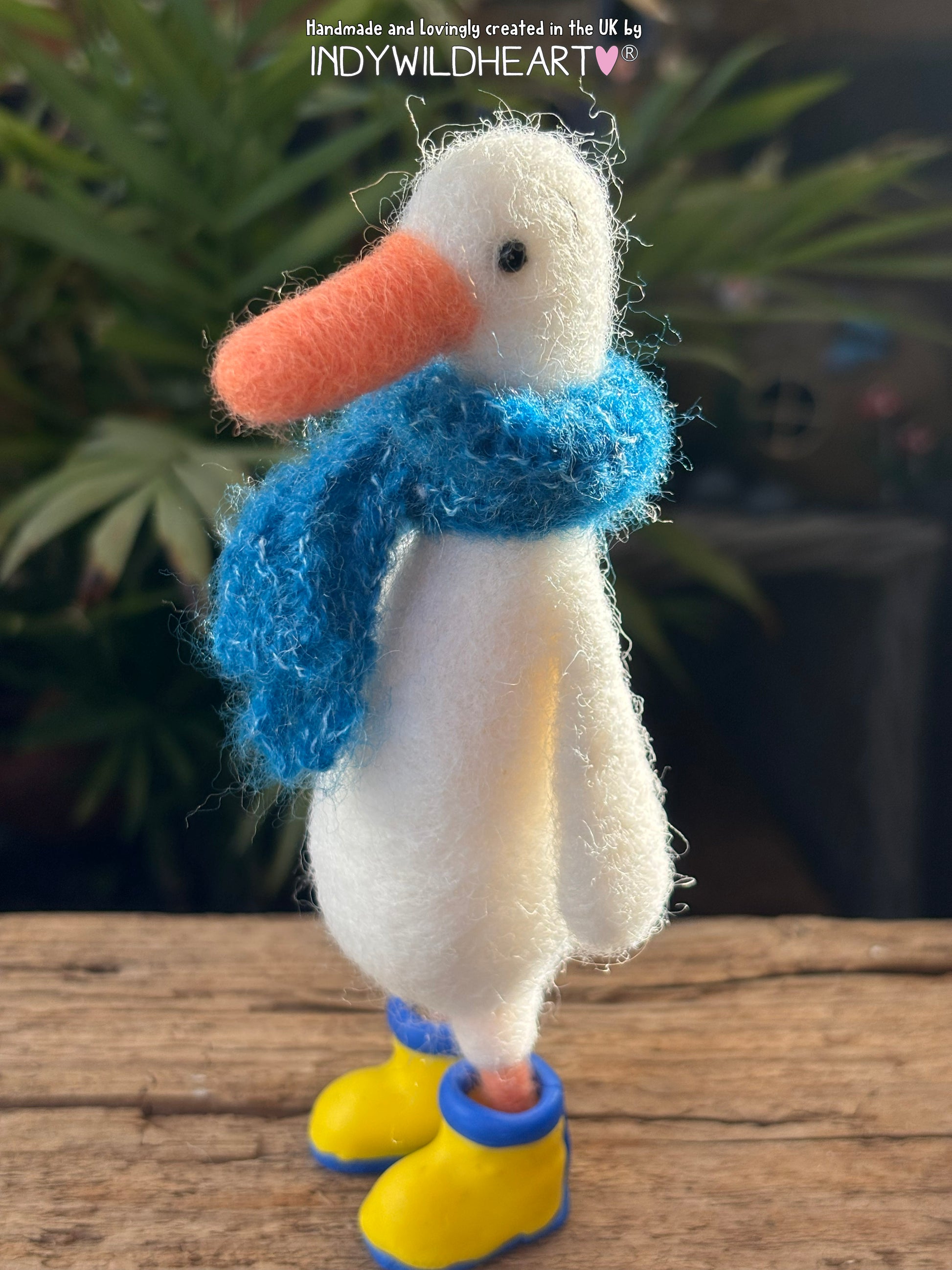 White duck needle felted  figurine with a blue scarf and yellow boots on a wooden surface.
