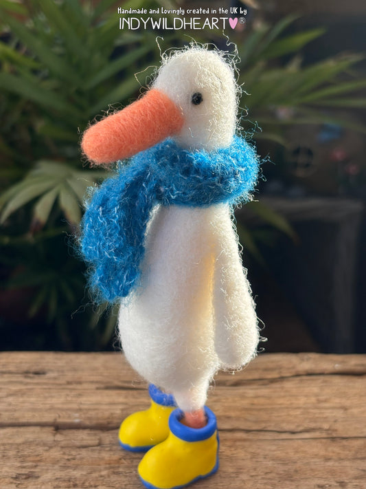 White duck needle felted  figurine with a blue scarf and yellow boots on a wooden surface.